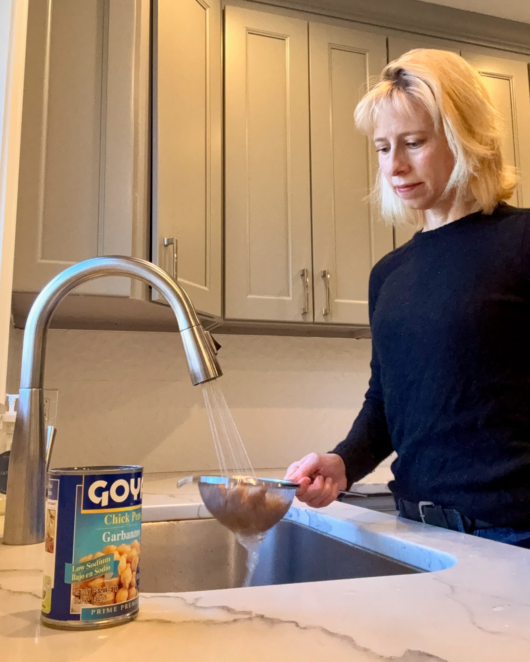 A woman rinses chickpeas in a strainer at a kitchen sink