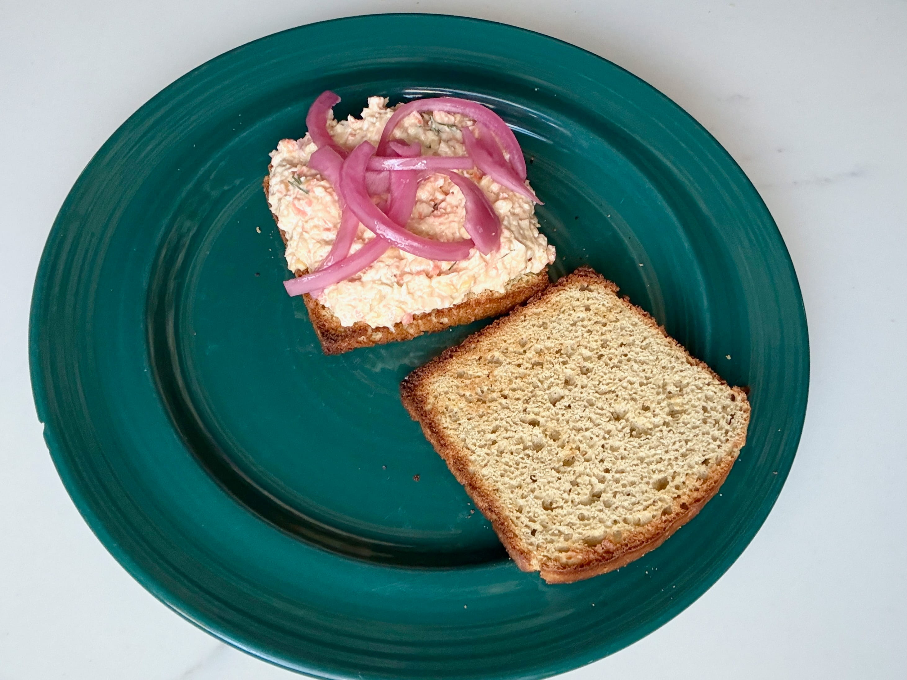 On a plate, the top slice of bread on a sandwich sits to the side, and the other slice is spread with chickpea tuna salad and pickled onions