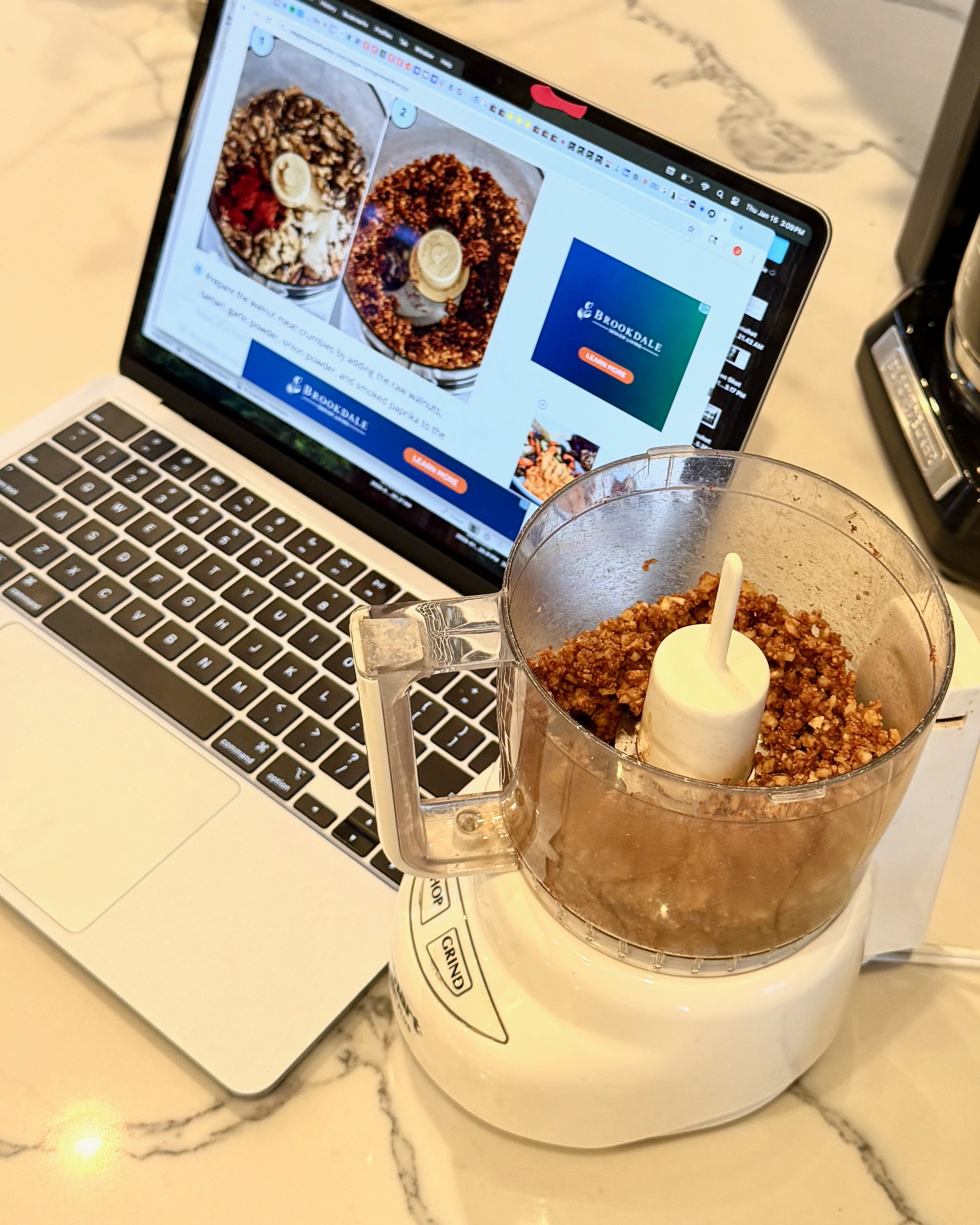 A laptop computer displays a recipe behind a food processor filled with a brown mixture