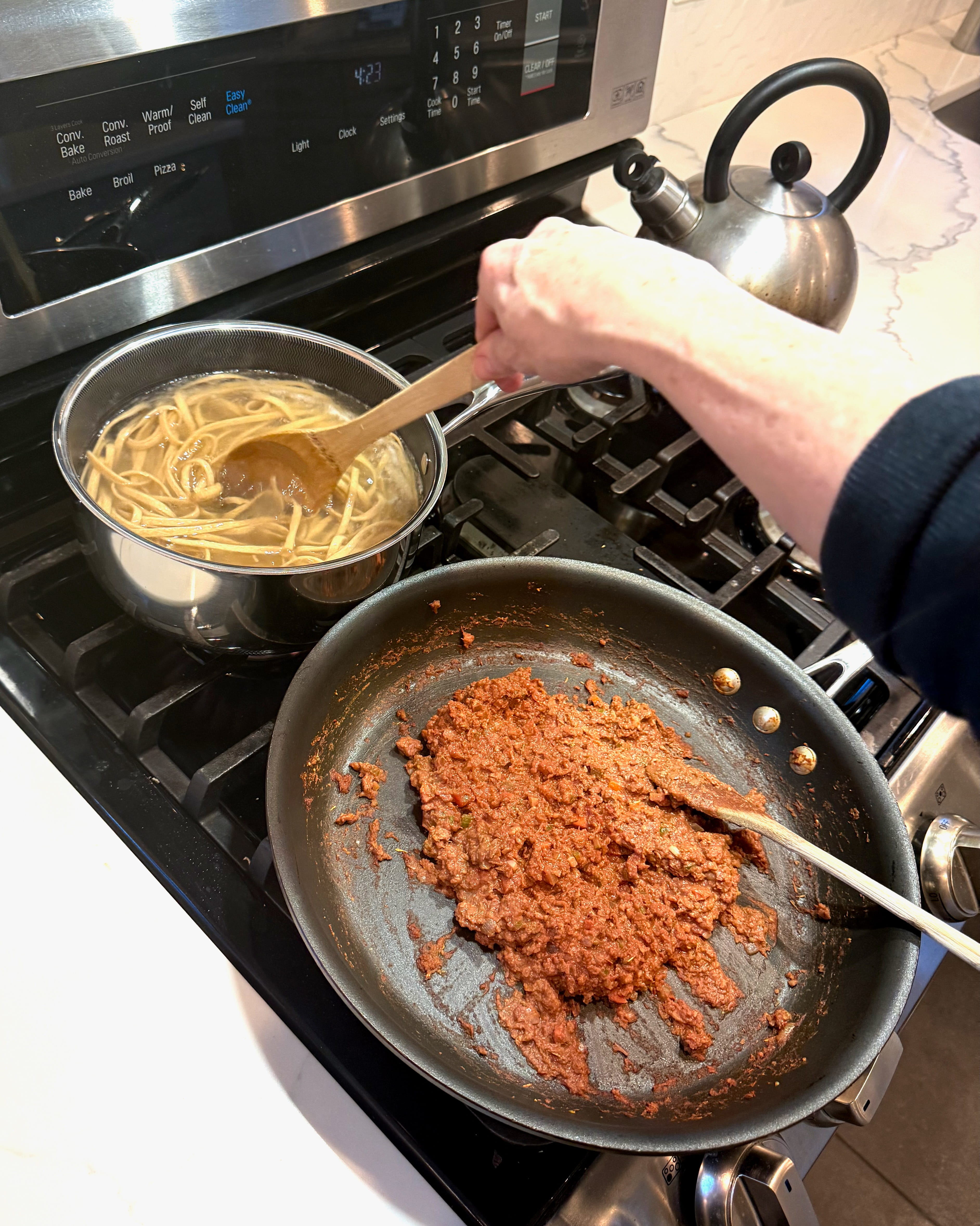 A person's hand stirs one of two pans of food cooking on a stove